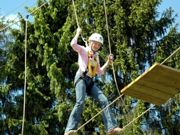 Hochseilgarten Dankern Kind mit Sicherheitsgurt balanciert auf hängenden Holzplattformen im Hochseilgarten vor Nadelbäumen