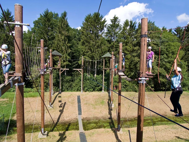 Menschen klettern mit Sicherungsausrüstung auf Holzelementen eines Hochseilgartens im Grünen.