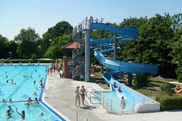 Freibad Hengstforde Das Freibad Hengstforde zeigt einen sonnigen Sommertag mit einem großen Schwimmbecken, das voller fröhlicher Menschen ist, umgeben von sattgrünen Bäumen. Eine blaue Wasserrutsche schlängelt sich neben dem Becken hinunter, während Menschen entspannt auf Liegestühlen liegen oder im kristallklaren Wasser schwimmen.
