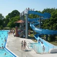 Freibad Hengstforde Das Freibad Hengstforde zeigt einen sonnigen Sommertag mit einem großen Schwimmbecken, das voller fröhlicher Menschen ist, umgeben von sattgrünen Bäumen. Eine blaue Wasserrutsche schlängelt sich neben dem Becken hinunter, während Menschen entspannt auf Liegestühlen liegen oder im kristallklaren Wasser schwimmen.