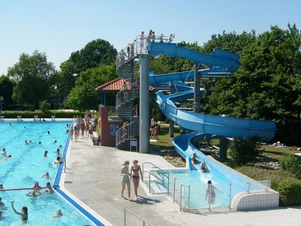 Freibad Hengstforde Das Freibad Hengstforde zeigt einen sonnigen Sommertag mit einem großen Schwimmbecken, das voller fröhlicher Menschen ist, umgeben von sattgrünen Bäumen. Eine blaue Wasserrutsche schlängelt sich neben dem Becken hinunter, während Menschen entspannt auf Liegestühlen liegen oder im kristallklaren Wasser schwimmen.