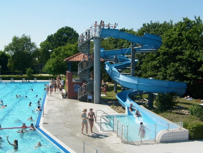 Freibad Hengstforde Das Freibad Hengstforde zeigt einen sonnigen Sommertag mit einem großen Schwimmbecken, das voller fröhlicher Menschen ist, umgeben von sattgrünen Bäumen. Eine blaue Wasserrutsche schlängelt sich neben dem Becken hinunter, während Menschen entspannt auf Liegestühlen liegen oder im kristallklaren Wasser schwimmen.
