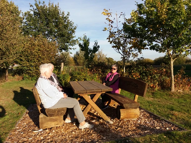 Gäste auf dem Rastplatz Hengstforde Zwei ältere Personen und eine jüngere Frau sitzen entspannt an einem Holztisch auf dem Rastplatz Hengstforde, umgeben von Bäumen und Wiesen im warmen Sonnenlicht.