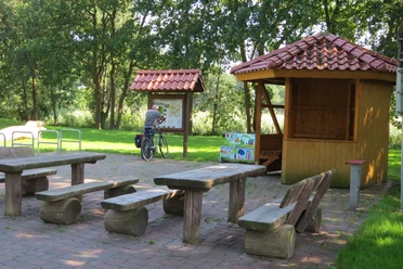 Rastplatz Am Drakamp Holzpicknicktische stehen auf gepflastertem Gelände, flankiert von einer Info-Tafel und einem Radfahrer.