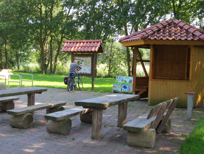 Rastplatz Am Drakamp Holzpicknicktische stehen auf gepflastertem Gelände, flankiert von einer Info-Tafel und einem Radfahrer.