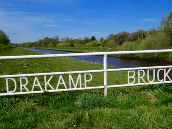 Drakamp Brücke Das Bild zeigt das leuchtende Schild "Drakamp Brücke" vor einem grasbewachsenen Ufer und einem ruhigen Flusslauf unter wolkenfreiem Himmel. Der Kontrast zwischen dem weißen Geländer und der satten grünen Vegetation vermittelt Ruhe und Gelassenheit. Der Horizont ist durch zart grüne Baumkronen gesäumt, die einen malerischen Übergang zum Himmel bilden. Dieses Szenario strahlt eine harmonische, ländliche Idylle aus.