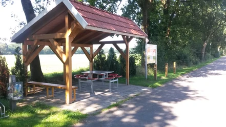 Rastplatz Alter Kirchweg Ein hölzerner Unterstand mit rotem Ziegeldach bietet Schutz, umgeben von Bäumen und neben einem Weg.