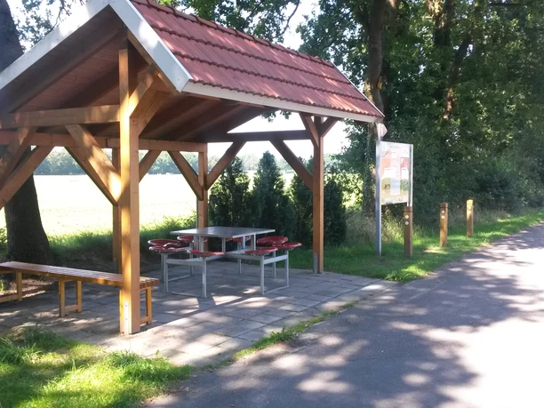 Rastplatz Alter Kirchweg Ein hölzerner Unterstand mit rotem Ziegeldach bietet Schutz, umgeben von Bäumen und neben einem Weg.