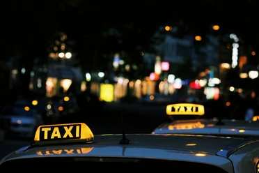 taxi Im Bild sind zwei Taxis mit leuchtendem Taxi-Schild auf dem Dach in einer belebten nächtlichen Straße zu sehen.