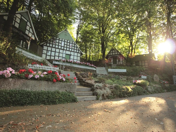 Freilichtbühne Kahle Wart Freilichtbühne umgeben von Fachwerkhäusern im Wald mit bunten Blumen und Sonnenlicht im Hintergrund.