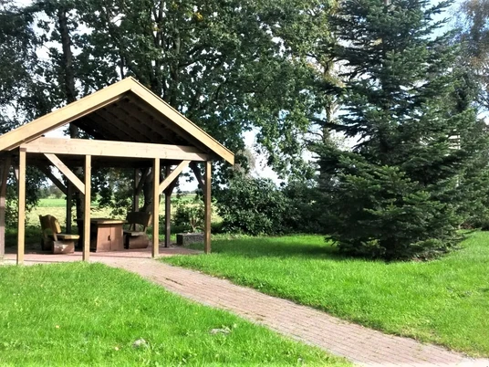 Rastplatz Bokelermoor Ein überdachter Rastplatz auf grüner Wiese, umgeben von Bäumen, mit einem gepflasterten Weg.