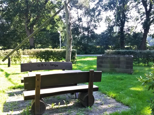 Rastplatz Tange Ein Rastplatz mit einer dunklen Holzbank und einem Tisch, umgeben von grüner Wiese und Bäumen.