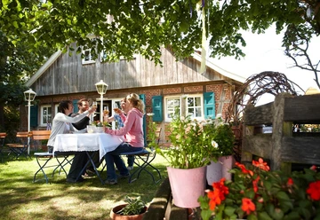 Radfahren im Emsland Vier Personen sitzen im Garten eines Landhauses und stoßen bei einer Mahlzeit unter Bäumen an.