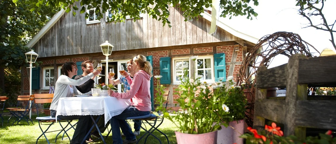 Radfahren im Emsland Vier Personen sitzen im Garten eines Landhauses und stoßen bei einer Mahlzeit unter Bäumen an.