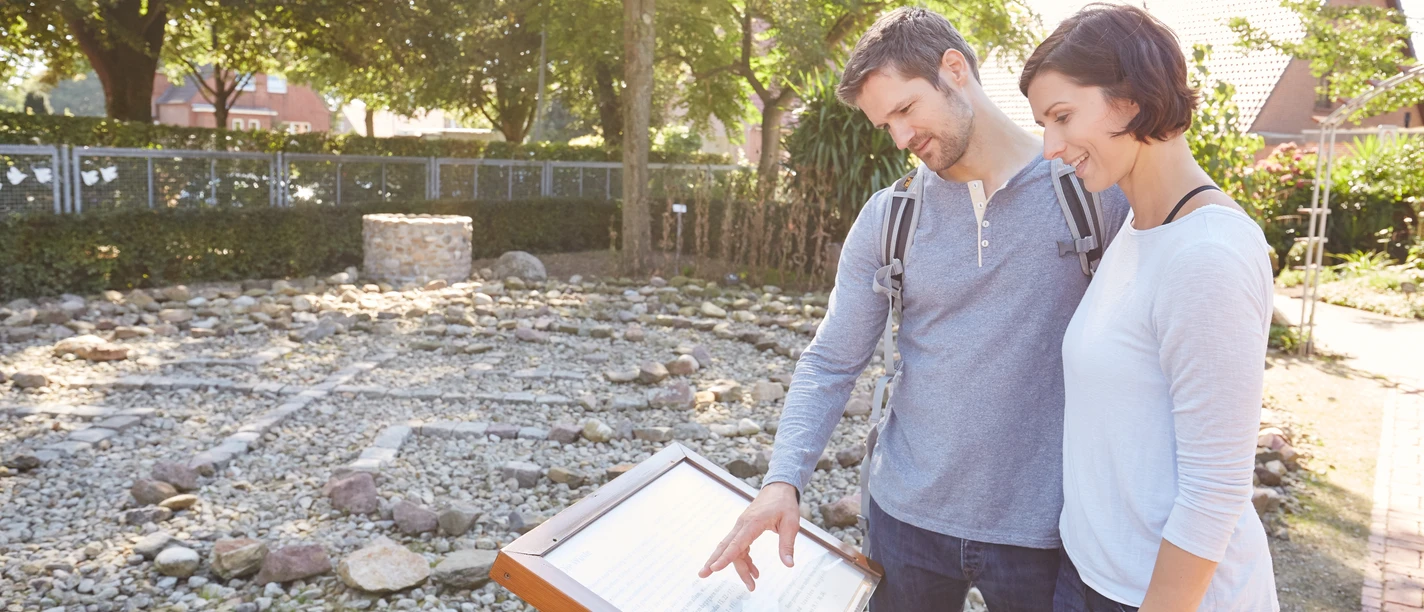 Bibelgarten Werlte Zwei Personen betrachten eine Informationstafel im Bibelgarten Werlte bei sonnigem Wetter.