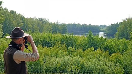 Een persoon met een hoed bekijkt door een verrekijker een groen merenlandschap in het Geester Wasserreich.