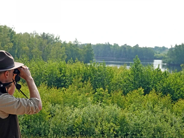 Een persoon met een hoed bekijkt door een verrekijker een groen merenlandschap in het Geester Wasserreich.
