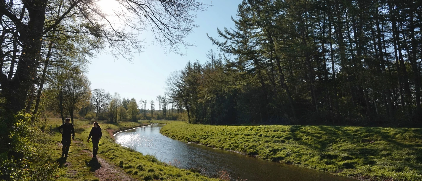 Wanderung entlang der Nordradde Zwei Wandernde folgen einem sonnenbeschienenen Pfad entlang eines ruhig fließenden Flusslaufs.