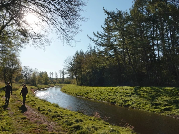 Wanderung entlang der Nordradde Zwei Wandernde folgen einem sonnenbeschienenen Pfad entlang eines ruhig fließenden Flusslaufs.