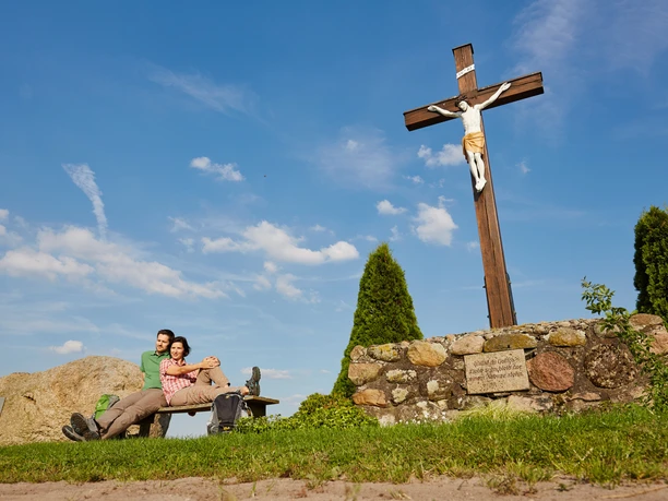 Ubben Krüss Zwei Wandernde ruhen auf einer Bank neben einem großen Holzkreuz unter blauem Himmel.