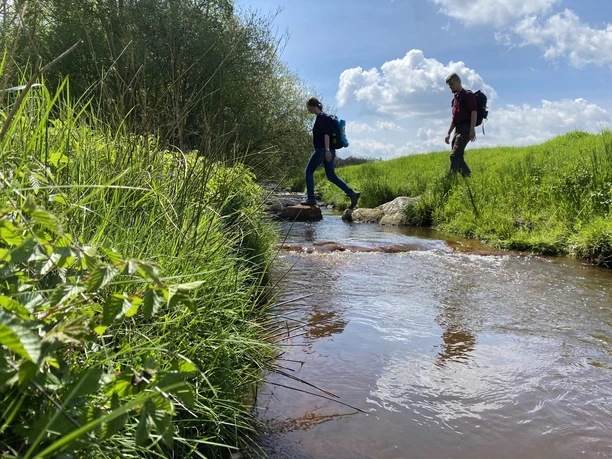 Oheüberquerung Zwei Wandernde überqueren auf Steinen einen Bach im grünen Ohetal unter blauem Himmel.