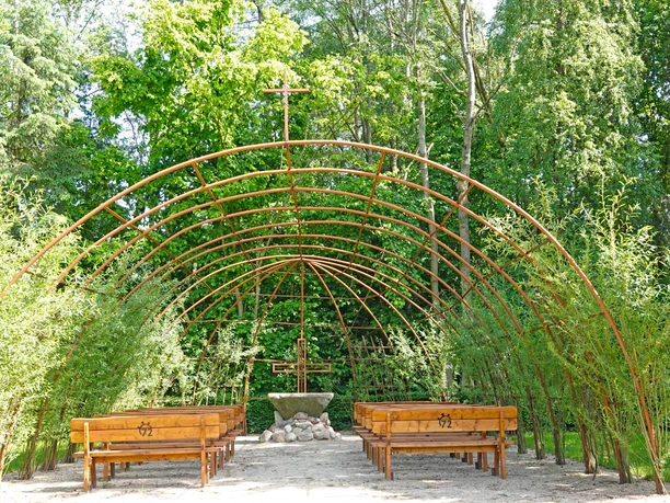 Weidenkirche Bruneforth Weidenkirche mit offenen Rutenbögen, Holzbänken und Altar, umgeben von grünem Laub im Freien