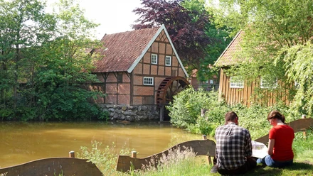 Historische Wassermühle mit Fachwerkhaus am Teich, umgeben von Bäumen, zwei Menschen im Vordergrund.