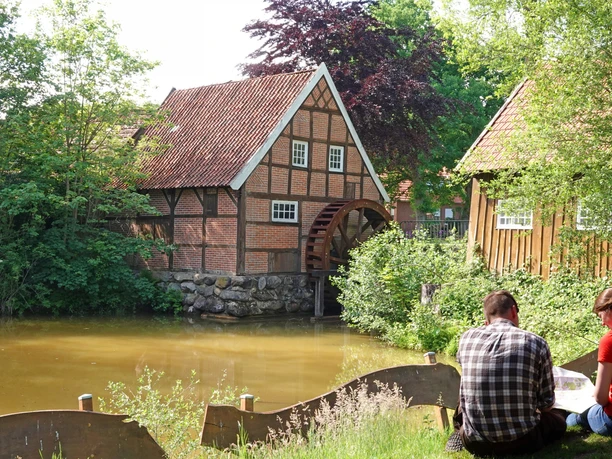 Historische Wassermühle mit Fachwerkhaus am Teich, umgeben von Bäumen, zwei Menschen im Vordergrund.