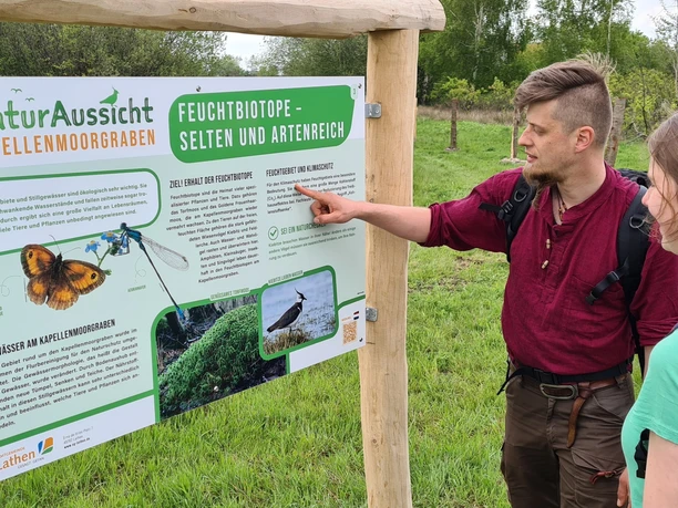 Zwei Wandernde betrachten eine Infotafel zu Feuchtbiotopen am Kapellenmoorgraben in Niederlangen.