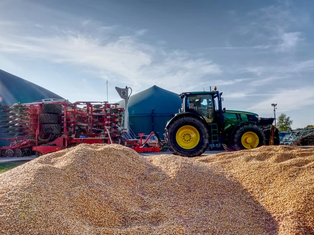Fahrzeug Hof Engelken Grüner Traktor mit roter Landmaschine vor Silos und Getreidehaufen unter blauem Himmel im Sonnenschein.