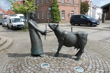 Pastor sine Koh Bronzeskulptur eines Pastors mit einer Kuh auf gepflastertem Platz vor rot gemauerten Gebäuden.