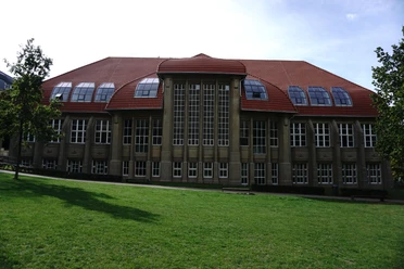 Historisches Backsteingebäude mit gewölbtem Dach und großen Fenstern vor grüner Wiese.