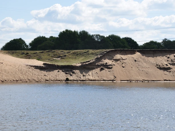 Meppener Kuhweide Sandböschung mit Seeufer bei Meppen, umgeben von Grasflächen und Bäumen unter Wolkenhimmel.