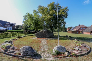 Opferstein Börger Großer Findling im Grünen, von Bäumen umgeben, eingerahmt von Steinen und Rasenfläche im Dorf.