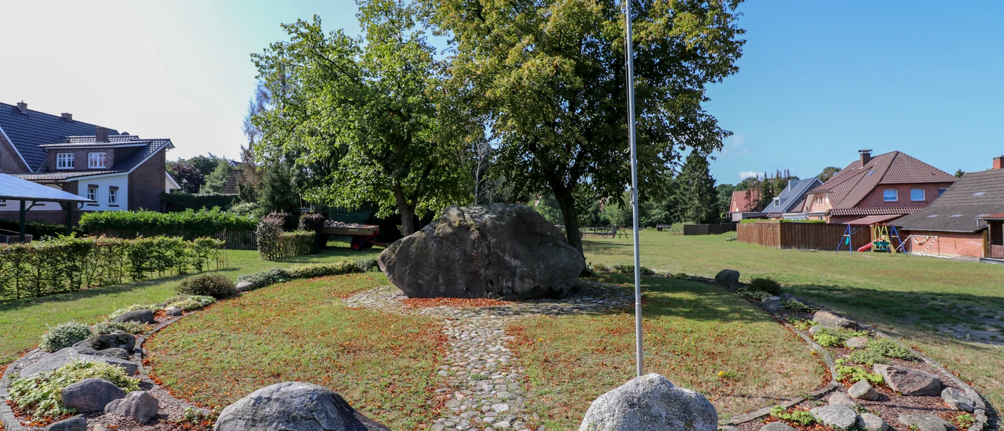 Opferstein Börger Großer Findling im Grünen, von Bäumen umgeben, eingerahmt von Steinen und Rasenfläche im Dorf.