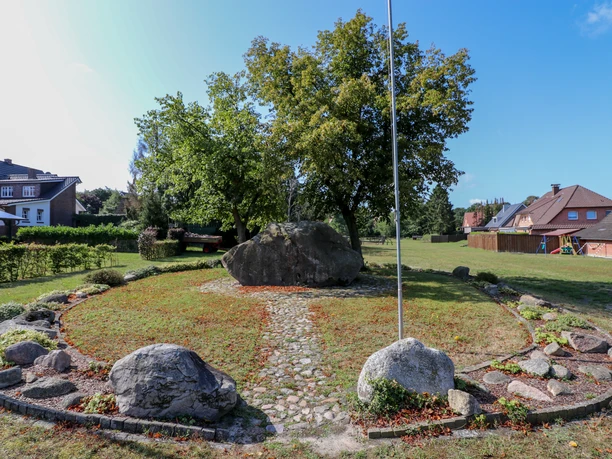 Opferstein Börger Großer Findling im Grünen, von Bäumen umgeben, eingerahmt von Steinen und Rasenfläche im Dorf.
