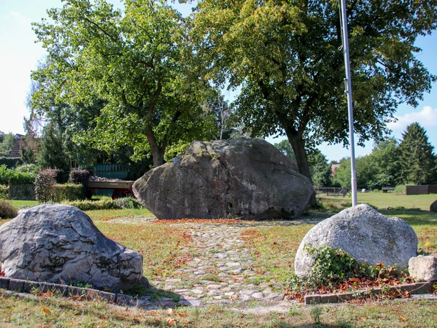 Opferstein Börger Großer Findling im Grünen, von Bäumen umgeben, mit gepflastertem Weg und weiteren Steinen im Vordergrund.