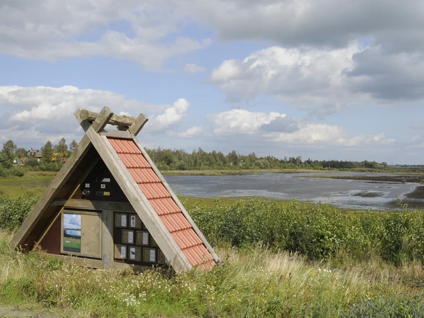 Naturschutzgebiet Rühler Moor Holzunterstand mit rotem Dach am Feuchtgebiet, dahinter weite Moorlandschaft unter Wolkenhimmel.