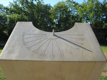 Steinerne Sonnenuhr in einem grünen Park, umgeben von Bäumen, zeigt römische Ziffern zur Zeitmessung.