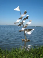 Eine moderne, silberne Skulptur eines Segels mit Wellen steht im klaren Wasser eines Sees.