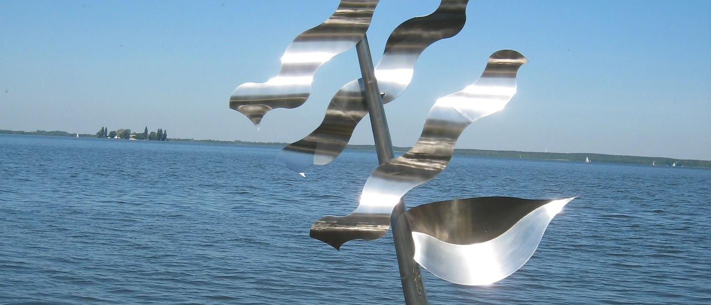 Eine moderne, silberne Skulptur eines Segels mit Wellen steht im klaren Wasser eines Sees.