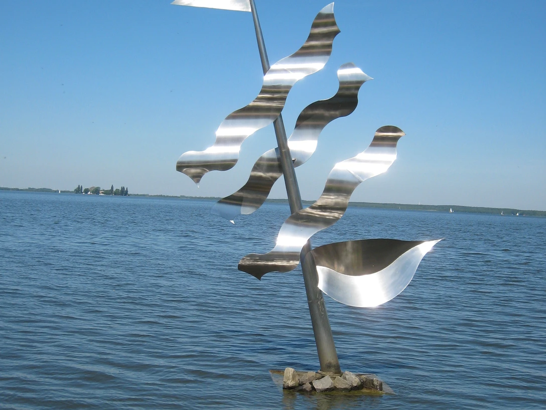 Undines Traum Eine moderne, silberne Skulptur eines Segels mit Wellen steht im klaren Wasser eines Sees.
