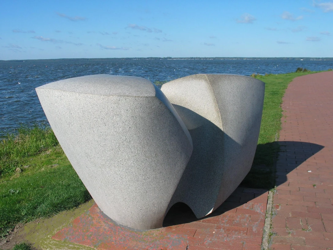 Moderne Skulptur aus Stein, bestehend aus geschwungenen Formen, steht vor einem See mit blauem Himmel.
