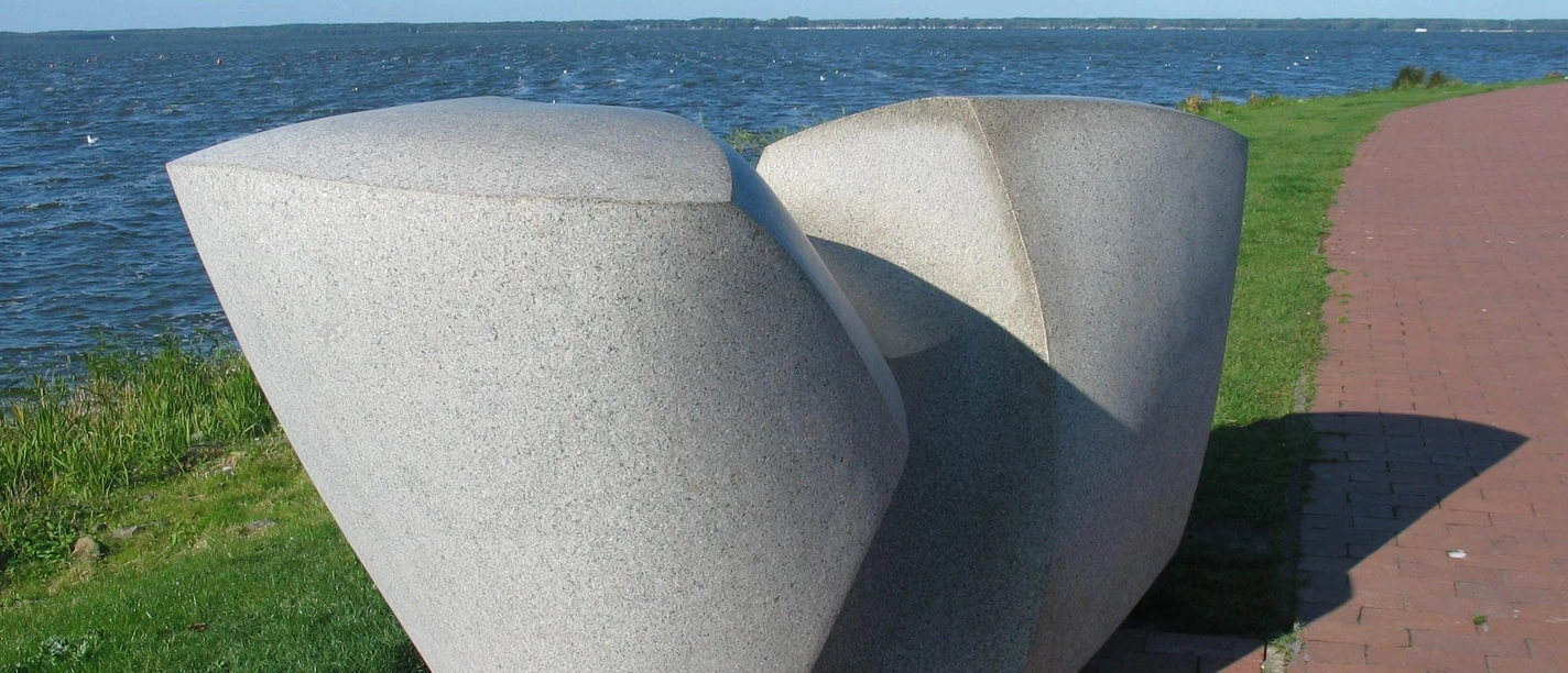 Swingende Welle Moderne Skulptur aus Stein, bestehend aus geschwungenen Formen, steht vor einem See mit blauem Himmel.