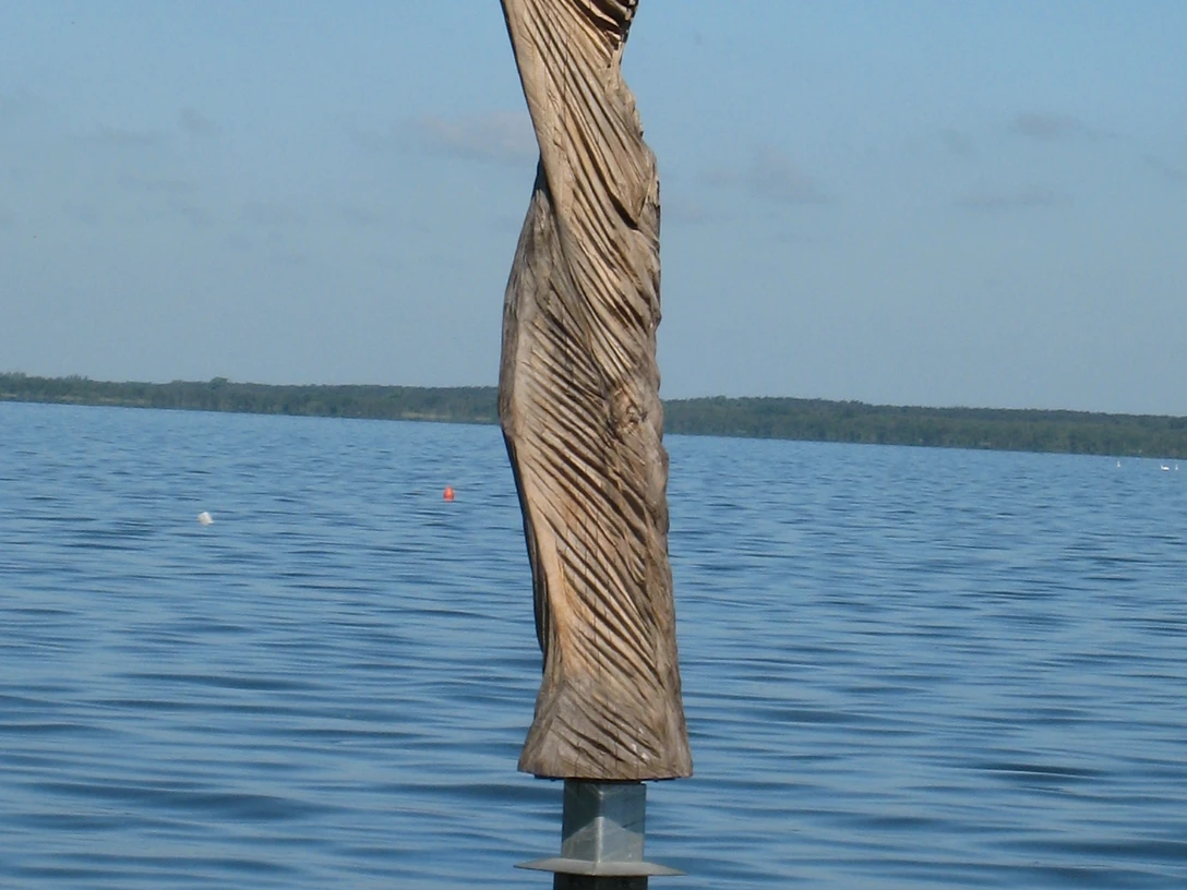 Skulptur aus Holz im Wasser, mit kunstvollen Falten, die dem Wind nachempfunden sind, vor blauem Himmel.