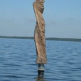 Skulptur aus Holz im Wasser, mit kunstvollen Falten, die dem Wind nachempfunden sind, vor blauem Himmel.