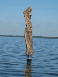 Papagena im Wind Skulptur aus Holz im Wasser, mit kunstvollen Falten, die dem Wind nachempfunden sind, vor blauem Himmel.