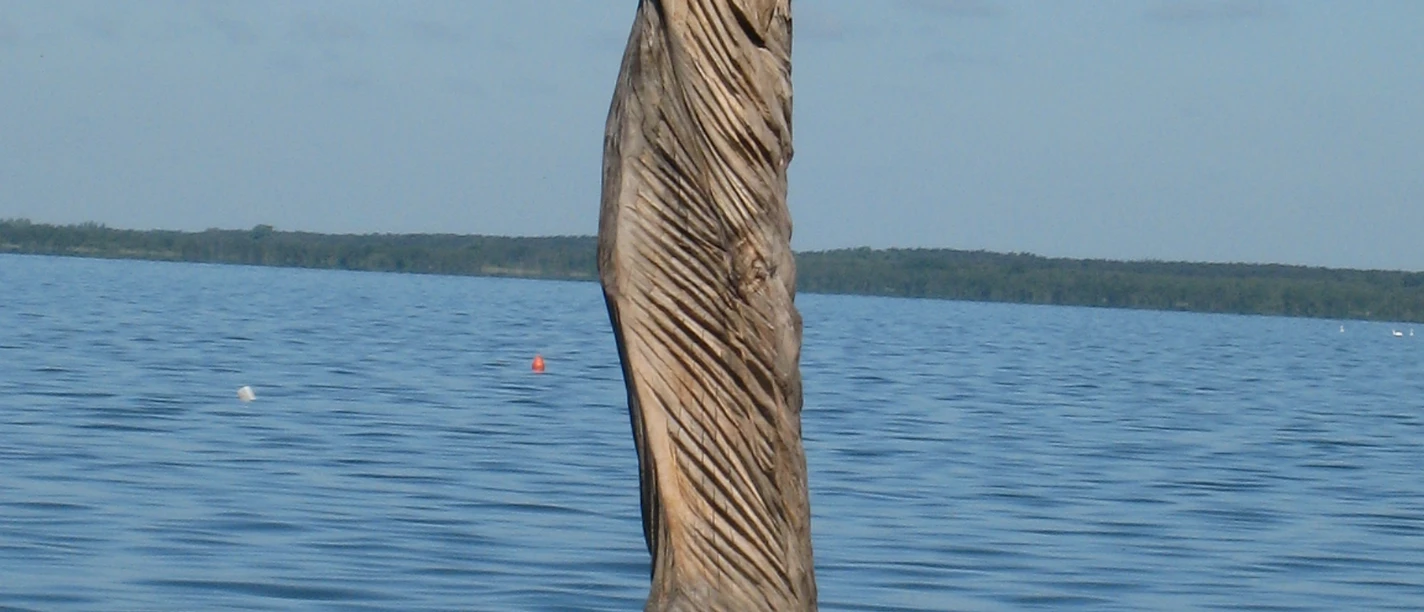 Papagena im Wind Skulptur aus Holz im Wasser, mit kunstvollen Falten, die dem Wind nachempfunden sind, vor blauem Himmel.