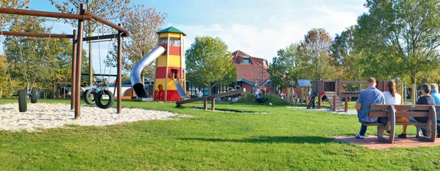 Abenteuerspielplatz Greetsiel Kinder spielen auf einer großen Wiese mit Holzschaukeln, Rutsche und rot-gelbem Aussichtsturm in Greetsiel.