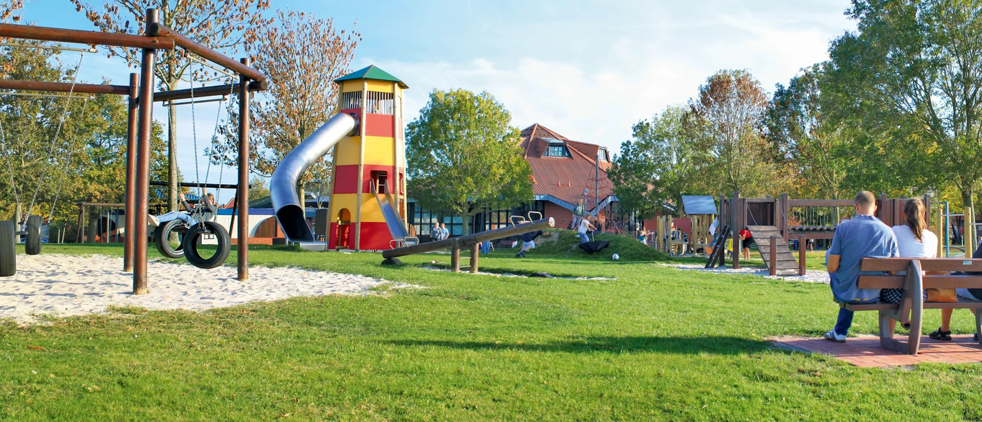 Abenteuerspielplatz Greetsiel Kinder spielen auf einer großen Wiese mit Holzschaukeln, Rutsche und rot-gelbem Aussichtsturm in Greetsiel.